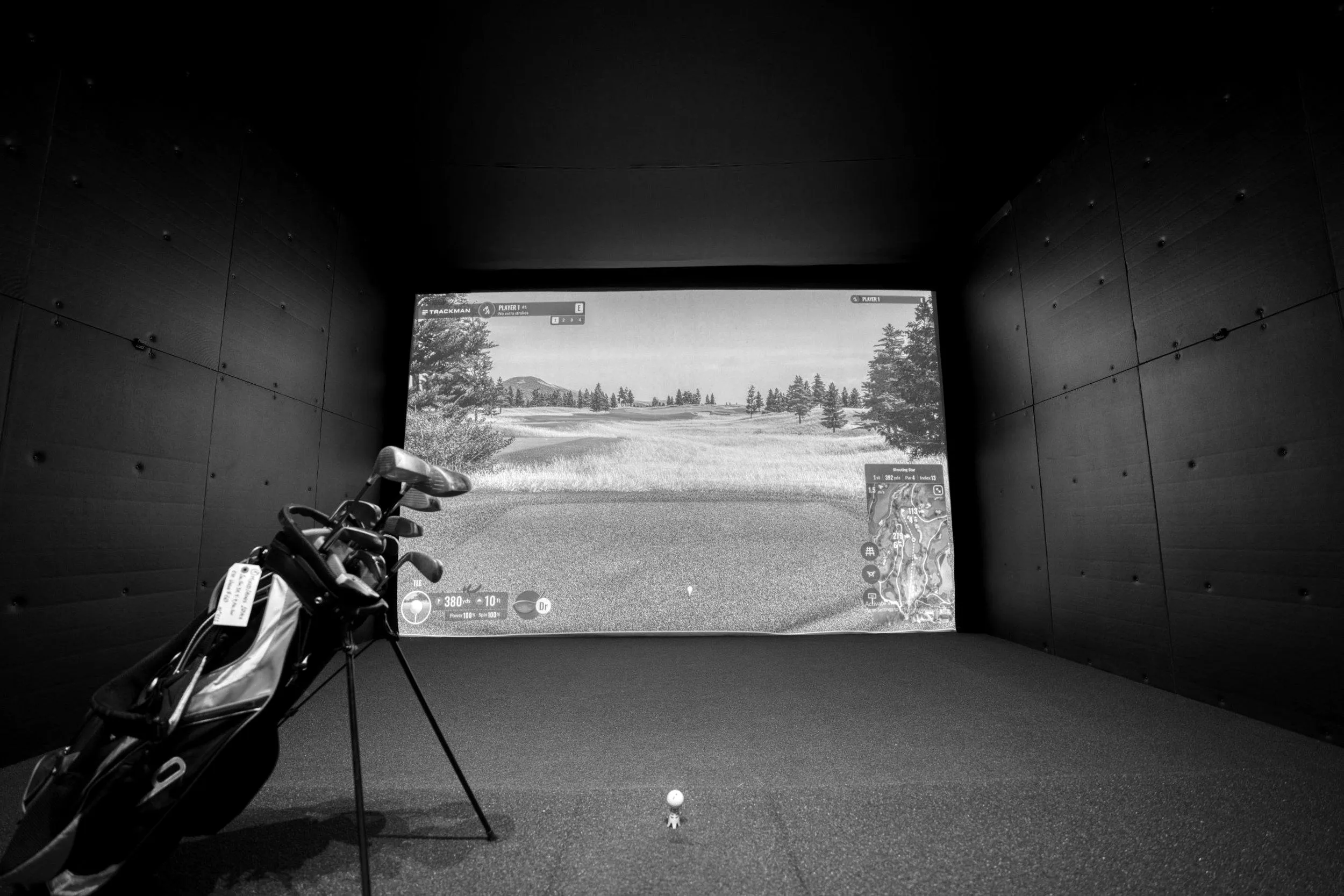 Svartvit bild av ett simulatorrum med impact screen, slagmatta och en golfbag, och en golfboll på en simulatorpegg